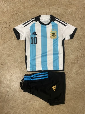 Argentina Messi fotbollströja och shorts - Säljer ett Argentina fotbollskit med Messi #10. Tröjan har blå och vita ränder, svarta detaljer och guldigt AFA-emblem. Shortsen är svarta med blå ränder och Adidas-logga i guld. Perfekt för dig som älskar fotboll och vill köra stilen på planen.