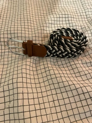 Svart och vit flätad bälte - Snyggt flätat bälte i svart och vitt med bruna skinn-detaljer vid spännet och änden. Spännet är i silverfärgad metall och bältet har en cool, randig look som passar till jeans eller chinos. Materialet är mjukt och flexibelt, vilket gör det bekvämt att bära hela dagen.