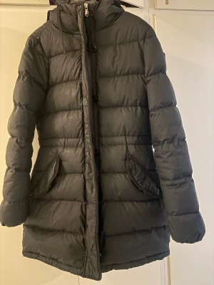 Svart pufferjacka från GANT, S - Säljer en svart pufferjacka från GANT i storlek S. Jackan har huva, dragkedja och knappar framtill samt två fickor med lock. Midjan är justerbar! Den är quiltad och har en längre modell som håller dig varm under kalla dagar. Perfekt för dig som gillar stilren och funktionell design.