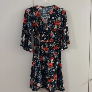 Blommig omlottklänning från Mango - Supersnygg omlottklänning från Mango med blommigt mönster i rött, blått och vitt på mörk botten. Klänningen har v-ringning, trekvartsärmar och knyt i midjan. Perfekt för dig som gillar romantisk stil och vill ha något som sticker ut.
