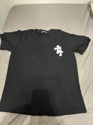 Svart t-shirt från Chrome Hearts - Svart t-shirt från Chrome Hearts med vit tryckt logga och korsmotiv både fram och bak. Klassisk rund hals och korta ärmar. Materialet är mjuk bomull som känns skön mot huden. Snygg och stilren design med tydliga street-vibes och kontrasterande detaljer.