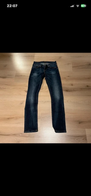 Mörkblå jeans från Nudie Jeans - Snygga mörkblå jeans från Nudie Jeans med klassisk femficksdesign och orangea kontrastsömmar. Modellen har rak passform och är tillverkad i slitstarkt bomullsmaterial. Bakfickorna har Nudie Jeans typiska broderade vågmönster.