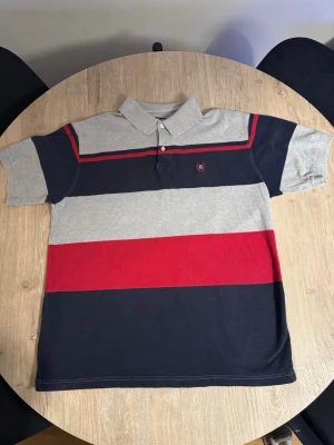 Randig pikétröja från Chaps Ralph Lauren - Säljer en pikétröja från Chaps Ralph Lauren i storlek XL. Tröjan har breda horisontella ränder i grått, marinblått och rött, med en broderad logga på bröstet. Klassisk krage och två knappar framtill. Tillverkad i 100% bomull för skön känsla.