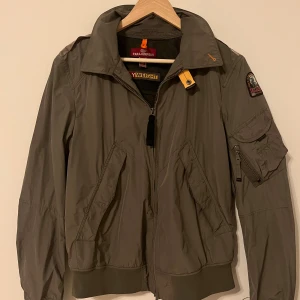 Olivgrön vindjacka från Parajumpers - Snygg olivgrön vindjacka från Parajumpers med PJS-märke på ärmen och gul detalj vid kragen. Jackan har dragkedja, flera fickor och ribbad nederkant. Perfekt för dig som gillar streetstyle och funktionella plagg. Litet osynligt hål finns med på bilden