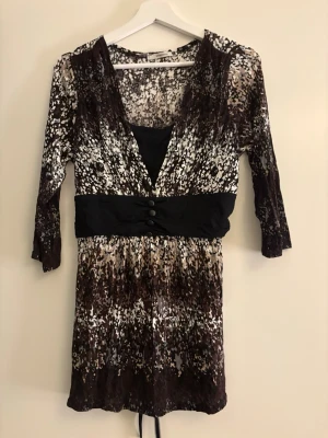 Mönstrad kortklänning från Wearhouse - Säljer en kortklänning från Wearhouse i storlek XL. Klänningen har ett brunt, svart och beige mönster, markerad midja med svarta knappar och v-ringad hals. Materialet är mjukt och stretchigt med 92% viskos och 8% elastan. Ärmarna är trekvarts och klänningen är tillverkad i Paris. Ej använd 
