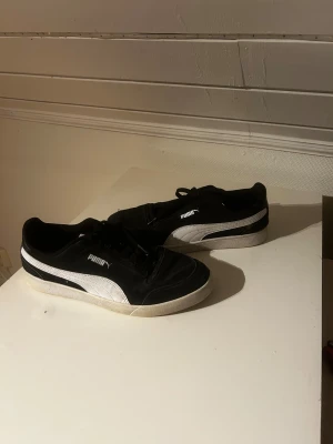 Svarta och vita sneakers från Puma - Säljer ett par snygga Puma sneakers i svart med vit sula och vit sidodetalj. Skorna har klassisk låg profil, rund tå och snörning. Materialet är en mix av syntet och skinn, med slitstark gummisula. Perfekt för dig som gillar stilrena och sportiga skor.