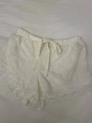Vita spets-shorts med snörning från ISABELla - Säljer ett par vita shorts från ISABEL i storlek S (36/38) , med vacker spets och dekorativ snörning framtill. De har högmidjad passform och fina spetsdetaljer längs kanterna som ger en romantisk vibe. Perfekta för varma dagar och chill häng.