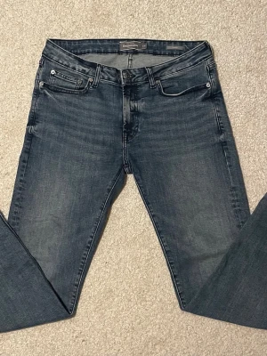 Jeans från Dressmann - Snygga blå slim fit jeans från Dressmann med klassisk femficksdesign och knappstängning. Jeansen har en stilren tvätt och raka ben, perfekta för dig som gillar en modern och enkel look. Materialet är slitstarkt jeans och passformen är slim.