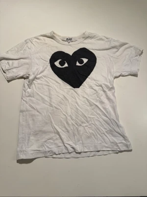 Vit t-shirt Comme des Garçons Play M - Säljer en vit t-shirt från Comme des Garçons Play i storlek M. Framsidan har ett stort svart hjärta med ögon, vilket ger en unik och cool vibe. T-shirten är kortärmad och gjord i mjuk bomull. Perfekt för dig som gillar streetwear och vill sticka ut.