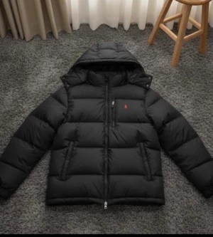 Svart pufferjacka från Polo Ralph Lauren - Svart pufferjacka från Polo Ralph Lauren med huva och diskret röd logga på bröstet. Jackan har två sidofickor, quiltad design och dragkedja framtill. Materialet är polyester och dun, perfekt för kalla dagar. Snygg och stilren modell som ger en cool vibe.