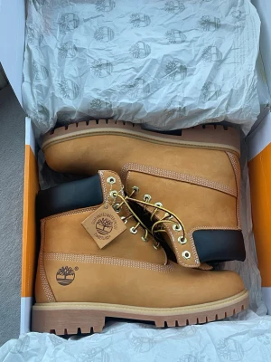 Timberland boots i beige skinn - Säljer ett par klassiska Timberland boots i beige skinn med svart vadderad krage och grov platt sula. Skorna har snörning med guldfärgade öljetter och tydlig Timberland-logga på sidan. Perfekta för höst och vinter, riktigt snygga till streetwear.