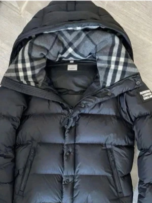 Svart pufferjacka från Burberry - Säljer en svart pufferjacka från Burberry med avtagbara ärmar, så den kan användas som väst. Jackan har rutigt foder i huvan, dragkedja och knappar framtill samt fickor med dragkedja. Märkespatch på ärmen och klassisk Burberry-design.