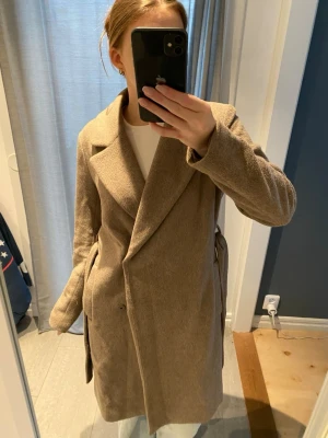 Beige kappa med bälte och knappar - Säljer en stilren beige kappa med bred krage och knappar framtill. Jackan har ett bälte i midjan och är helfodrad med lång ärm. Nypris runt 1800kr