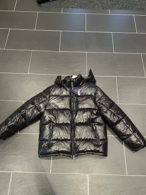 Svart pufferjacka från Polo Ralph Lauren - Svart pufferjacka från Polo Ralph Lauren med glansig finish och röd broderad logga på bröstet. Jackan har huva, två fickor med dragkedja och hel dragkedja framtill. Perfekt för kalla dagar och har en modern, boxig passform.