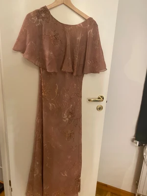 Lång blommig klänning med volang zara - Säljer en elegant långklänning från NA-KD i en varm beige ton med blommigt mönster. Klänningen har en vacker volang över axlarna och en öppen rygg. Materialet är lätt och känns som polyester, perfekt för sommarens tillfällen. Storlek L.