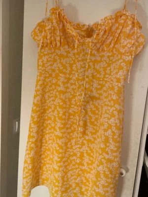 Gul blommig kortklänning Glamorous - Superfin gul klänning från Glamorous med vita blommor, volang vid halsen och smala axelband. Klänningen har en söt knytning framtill och dragkedja i ryggen. Perfekt för soliga dagar och sommarhäng.aldrig använt 