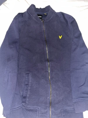 Mörkblå bomberjacka från Lyle & Scott - Snygg mörkblå bomberjacka från Lyle & Scott med gul broderad logga på bröstet. Jackan har ribbade muddar, dragkedja framtill och två sidofickor. Perfekt för dig som vill ha en stilren och enkel jacka med sportig vibe.