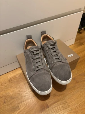 Grå mockasneakers från Christian Louboutin - Snygga grå sneakers i mocka från Christian Louboutin med klassisk röd sula och vita kanter. Skador på skorna det är därför priset är sänkt.