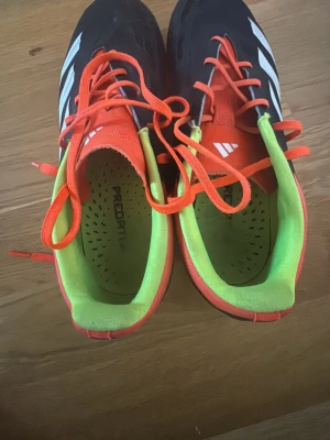 Adidas Predator fotbollsskor svart/orange - Säljer ett par Adidas Predator fotbollsskor med svart ovandel, vita ränder och orange detaljer. Skorna har snörning, rund tå och platt sula med dobbar för gräsplan. Insidan är limegrön och märkt med Predator. Perfekt för dig som vill ha grym kontroll på planen.