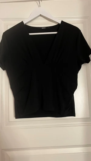 Svart v-ringad topp från Bik Bok - Säljer en svart topp från Bik Bok med djup v-ringning och korta ärmar. Toppen har en croppad passform och är tillverkad i mjukt syntetmaterial. Perfekt att styla med höga jeans eller kjol för en cool look.