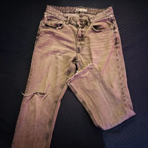 Grå ripped jeans från Zara, strl 42 - Snygga grå jeans från Zara i storlek 42 med slitna detaljer och hål vid knäet för en cool, avslappnad vibe. Klassisk femficksmodell med knappstängning och hög midja. Materialet är robust denim i bomull.