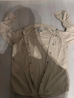 Beige overshirt från Jack & Jones - Snygg beige overshirt från Jack & Jones Core i slim fit. Jackan har knappar hela vägen, två bröstfickor och klassisk krage. Materialet är bomull och passar perfekt för lager-på-lager. Enkel och stilren design som funkar till många outfits. Saknar en knapp vid ärmen 
