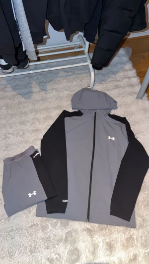 Under Armour träningsset grå/svart M - Snyggt träningsset från Under Armour i storlek M dessutom Limited Editon, består av en grå zip-hoodie med svarta ärmar och matchande grå byxor med svarta detaljer. Setet har vita loggor och dragkedja på jackan. Perfekt för gymmet eller löprundan.