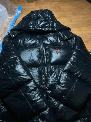 Svart pufferjacka från Polo Ralph Lauren - Svart glansig pufferjacka från Polo Ralph Lauren med huva och röd broderad logga på bröstet. Jackan har tvåvägsdragkedja, quiltad design och är tillverkad i syntetmaterial. Perfekt för kalla dagar och har en riktigt modern vibe.