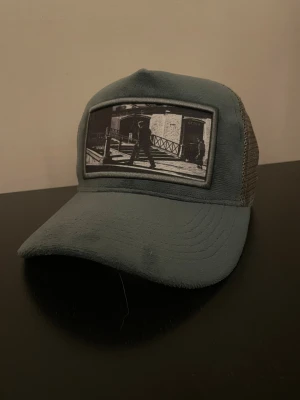 Grå keps från Emilio Pendenza - Säljer en grå keps från Emilio Pendenza med mesh på sidorna och baksidan. Framtill finns en svartvit patch med ett stadsmotiv. Kepsen har böjd skärm och justerbar passform. Perfekt för dig som vill ha en cool och urban stil.