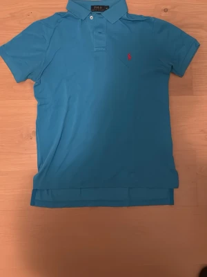 Blå pikétröja från Polo Ralph Lauren - Säljer en blå pikétröja från Polo Ralph Lauren i storlek M, men custom fit. Tröjan har klassisk krage, korta ärmar och knappar vid halsen. På bröstet finns det ikoniska röda broderade Polo-loggan. Perfekt för en chill och stilren look.