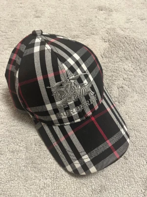 Burberry - Säljer en snygg Burberry keps med klassiskt rutigt mönster i svart, vitt och rött. Kepsen har broderad Burberry-logga framtill och justerbart spänne bak i metall. Perfekt accessoar för att lyfta din stil.