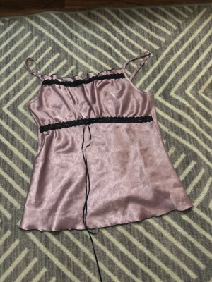 Ljuslila satin linnetopp med spets - Säljer en ljuslila linnetopp i satin med tunna axelband och svarta spetsdetaljer vid byst och midja. Toppen har en lätt glansig finish och en smickrande, loose passform. Storlek eller märke står ej på, men passar mig bra som är S.