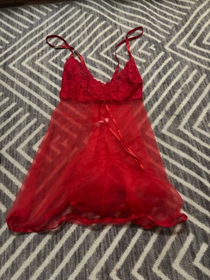 Röd spets babydoll med rosett - Söt babydoll i rött med spets upptill och tunna axelband. Transparent mesh i kjoldelen och en liten rosett framtill. Storleken står ej på plagget, men skulle säga att det är en M ungefär.