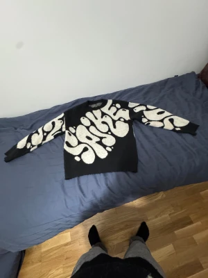 Svart stickad tröja med vit mönster - Säljer en svart stickad tröja med unikt vitt mönster som liknar abstrakta former eller ormar. Tröjan har rund hals och långa ärmar, perfekt för dig som gillar att sticka ut med cool design. Materialet är mjukt och ger en skön känsla.