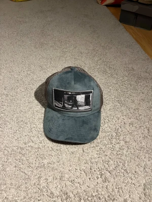 Grå meshkeps med tryck - Säljer en grå keps med mesh på baksidan och ett svartvitt tryck framtill som föreställer en stadsscen. Kepsen har böjd skärm och justerbar passform. Perfekt för dig som gillar streetwear och vill ha en cool accessoar.