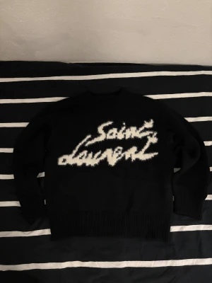 Svart Saint Laurent stickad tröja - Svart stickad tröja från Saint Laurent med vit, fluffig logotyp framtill. Tröjan har rund hals, ribbade muddar och lång ärm. Materialet känns mjukt och har en lyxig känsla, perfekt för dig som gillar statement-plagg med designerstil. Bara använd ett par gånger, fortfarande rikigt bra skick. Mycket varm o skön nu på vintern. Hör av dig om du har några frågor.