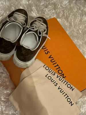 Louis Vuitton Time Out sneakers  - Säljer ett par snygga Louis Vuitton sneakers i mörkbrunt med klassiskt LV-monogram och vita detaljer. Skorna har snörning, rund tå och platt sula. Tillverkade i skinn med vit insida och kommer med originalkartong och dustbag. Skorna köptes i Nice, Frankrike 2021. Snörena och skorna kommer att tvättas/torkas av innan de skickas.