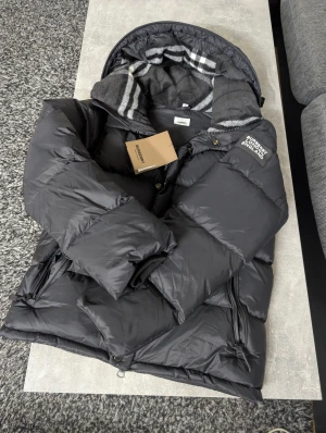 Burberry Puffer - Svart pufferjacka från Burberry med huva och klassiskt rutigt foder i grått och vitt. Jackan har dragkedjor på fickorna, snörning nedtill och en snygg Burberry-logga på ärmen. Perfekt för kalla dagar!