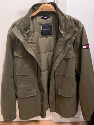 Olivgrön parkajacka Tommy Hilfiger - Snygg olivgrön parkajacka från Tommy Hilfiger med flera stora fickor framtill och klassisk logga på ärmen. Jackan har dragkedja och knappar, samt en hög krage. Perfekt för dig som gillar streetstyle och vill ha en jacka med coola detaljer.