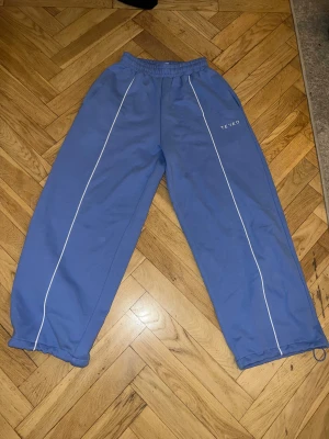 Blå wide leg joggers från TEVEO - Säljer ett par blå wide leg joggers från TEVEO med vita kontrastlinjer längs benen och elastisk midja. Byxorna har en loose passform och är tillverkade i mjuk bomull. Perfekta för en chill och avslappnad stil.