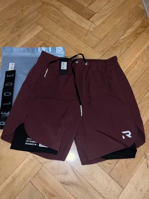 Burgundy träningsshorts från Relode - Säljer ett par burgundy träningsshorts från Relode med svart innerbyxa, snörning i midjan och vit logga på benet. Shortsen har en loose passform och är perfekta för gymmet eller löpning. Materialet är lätt och syntetiskt för maximal komfort.