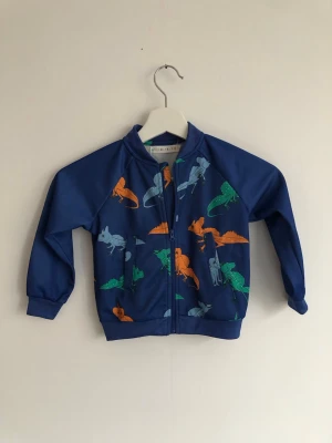 Färgglad tröja Minimarket - Färgglad jacka/tröja med ödlor på från minimarket. Fodret är varmt och mjukt. Storlek 86/92. Nyskick. Matchande byxor finnes och kan också säljas om så önskas(20 kr), men har några skrapor på knäna.