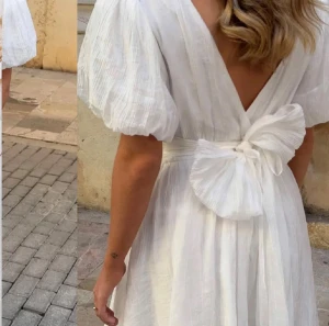 Vit kortklänning med puffärmar och rosett - Superfin vit kortklänning med puffiga kortärmar och djup v-ringning både fram och bak. Klänningen har en bred midja och en stor rosett i ryggen som knyts. Materialet är lätt och krinklat, vilket ger en luftig känsla. Perfekt för sommarens alla tillfällen!