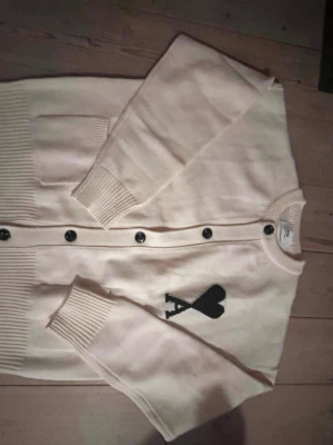 Vit kofta från Ami - Säljer en snygg vit kofta från Ami med svarta knappar och ett stort svart hjärta med bokstaven A på bröstet. Koftan är stickad, har långärmade och ribbade muddar samt nederkant. Perfekt för dig som gillar stilrena detaljer och vill ha något som sticker ut.