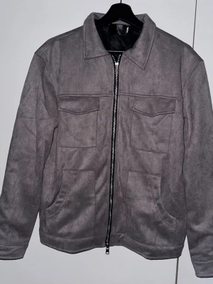 Grå mocka overshirt med dragkedja - Snygg grå overshirt i mocka-look med raka linjer och klassisk krage. Jackan har två stora bröstfickor med lock, sidofickor och en rejäl dragkedja framtill. Perfekt för lager-på-lager och ger en clean vibe till din outfit.