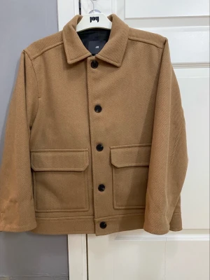 Beige jacka från H&M, storlek M - Snygg beige overshirt från H&M i storlek M. Jackan har klassisk krage, stora fickor framtill och svarta knappar. Materialet är mjukt och känns som ullblandning, perfekt för lager-på-lager. Passformen är normal och ärmarna är långärmade.
