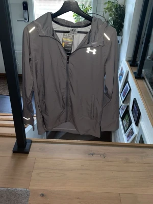 Grå vindjacka från Under Armour - Snygg grå vindjacka från Under Armour med huva och reflekterande detaljer. Jackan har dragkedja framtill, fickor på sidorna och logga på bröstet samt stor text på ryggen. Perfekt för träning utomhus när det blåser. Vid frågor är det bara att skriva😁