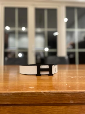 Svart och vit skinnväst med H-spänne - Säljer en stilren väst i svart och vitt med ett stort H-format spänne framtill. Västen är tillverkad i präglat skinn och har en klassisk, rak form. Perfekt för dig som gillar minimalistisk design och vill sticka ut med en snygg detalj.