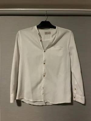 Vit henley-skjorta från Zara Boys - Vit henley-skjorta från Zara Boys i storlek 164. Skjortan har en strukturerad bomullsyta, knappar framtill och vid ärmslut, samt en liten bröstficka. Enkel och stilren design med lång ärm och rundad hals utan krage.