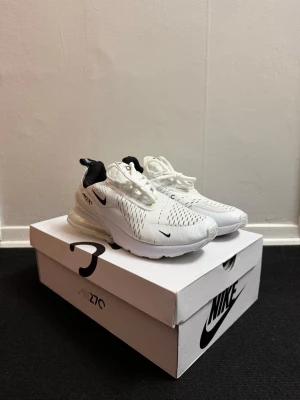 Nike Air Max 270 vita sneakers - Nike Air Max 270 sneakers i vit med svarta detaljer och synlig luftkudde i hälen. Ovandel i ventilerande syntetmaterial med snörning och rund tå. Snygg och modern design med svart logga på sidan och bakre flik för enkel påtagning.